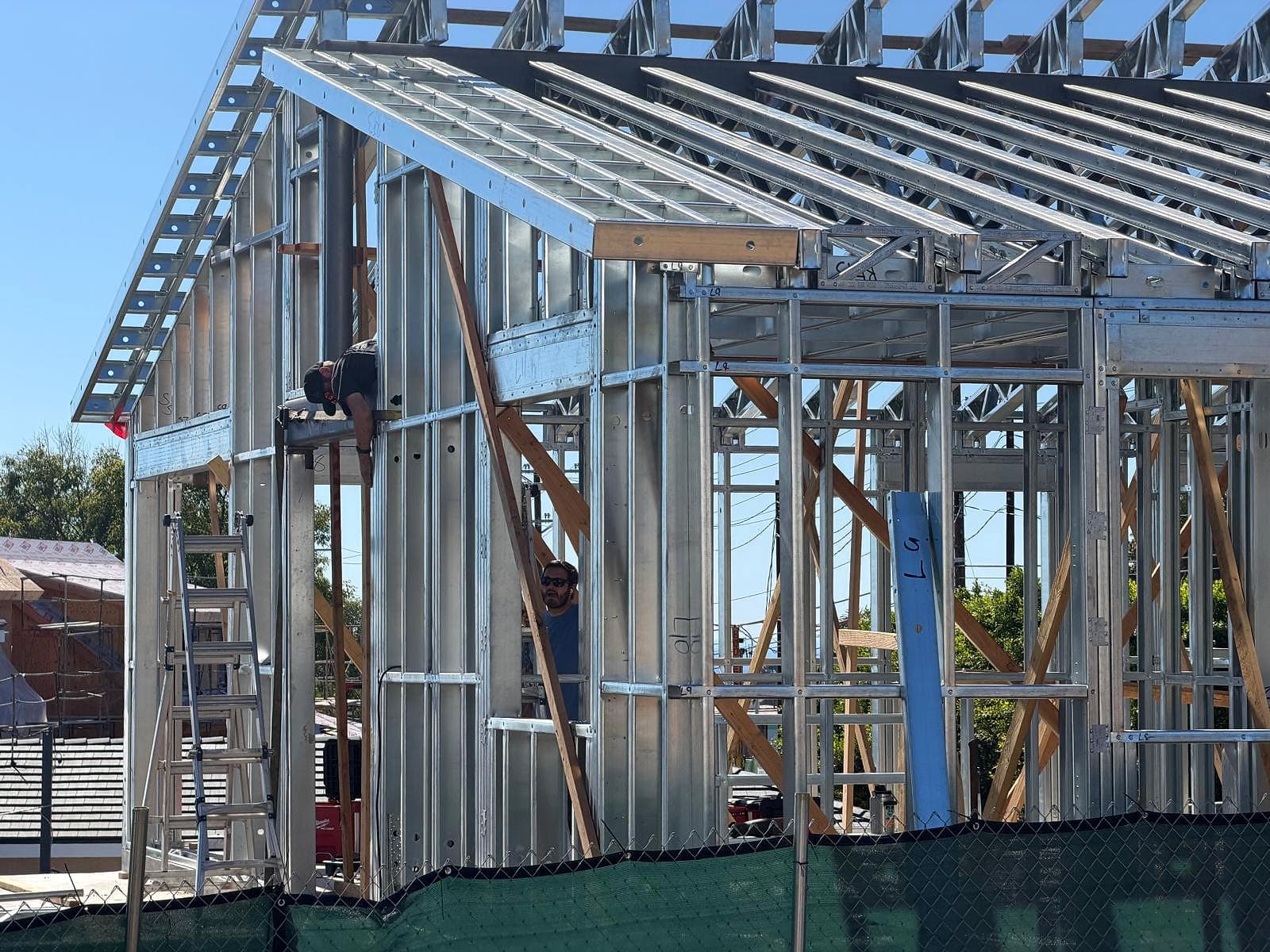 Two crew members working inside the steel wall framing under the roof, with a ladder-style steel eave overhang at the top left
