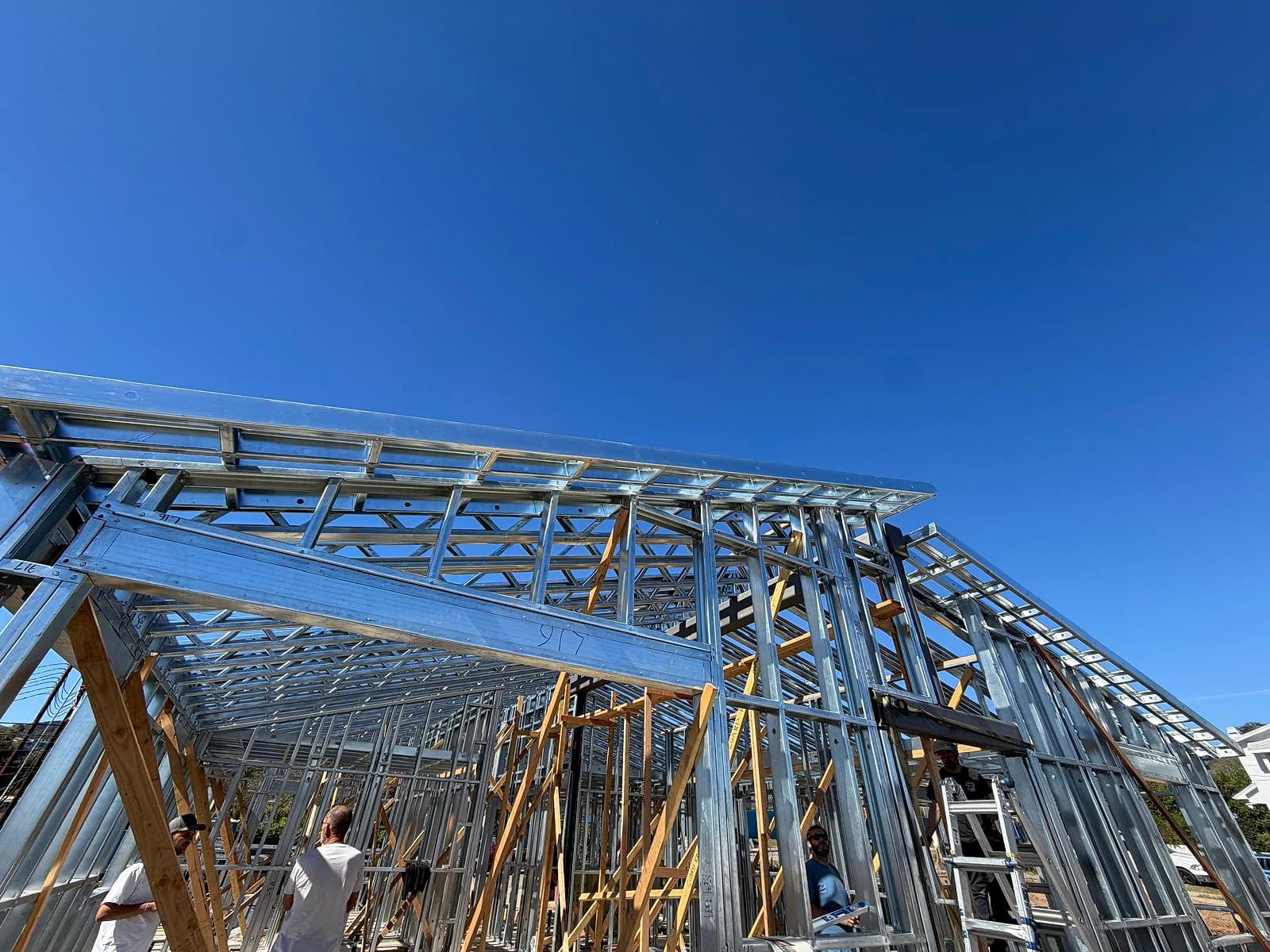 Looking up at steel roof framing and walls from the slab, crew members working below